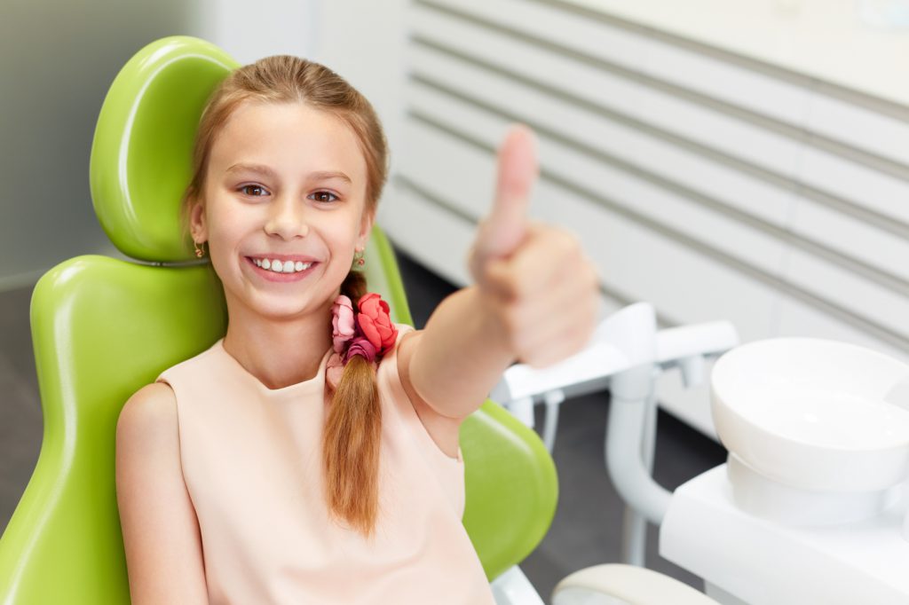 Portrait of happy girl shows thumb up gesture at dental clinic